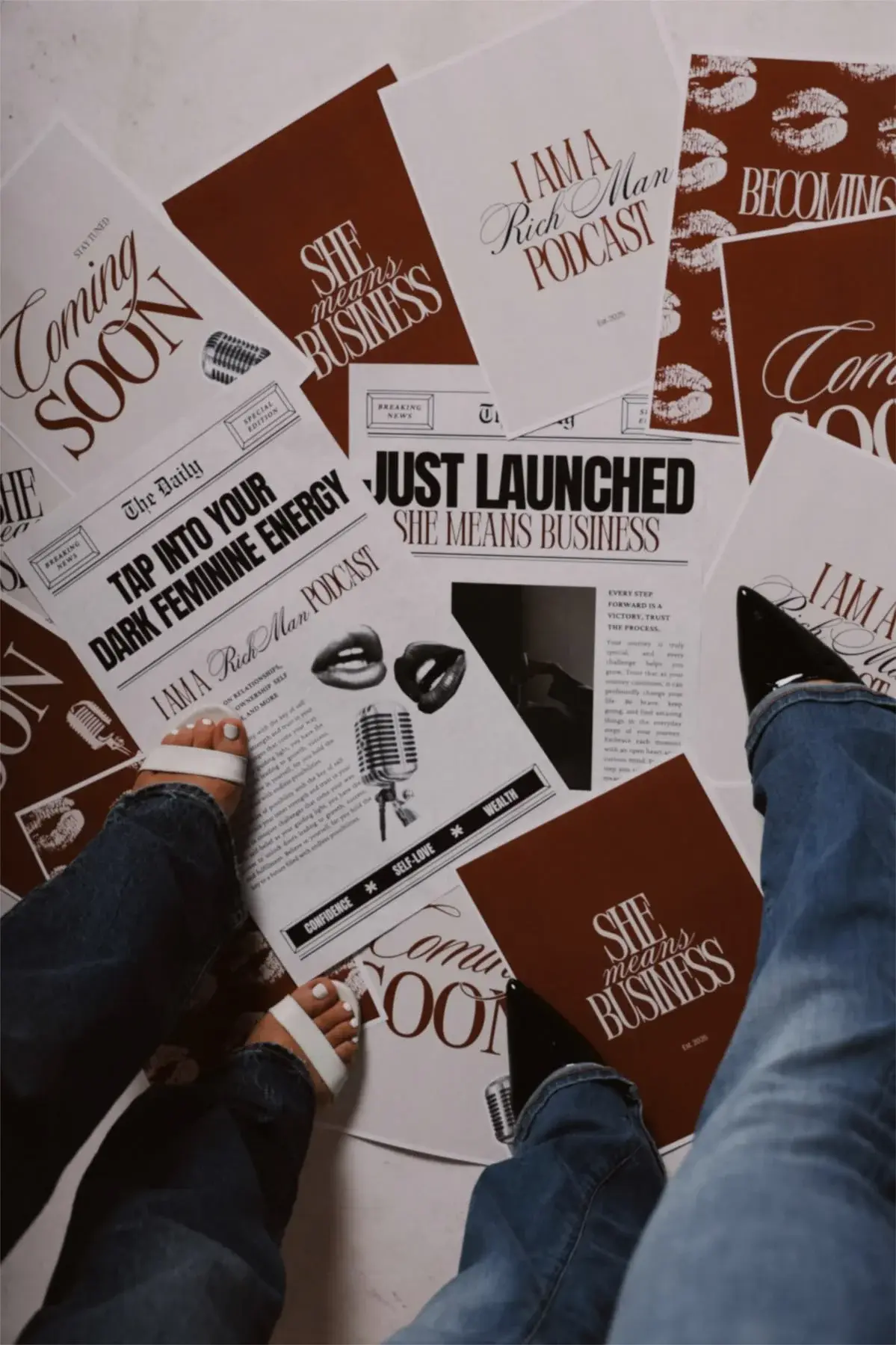 Top-down flat lay view of print advertisements, promotional flyers, and magazine layouts spread on the floor surrounded by two people.