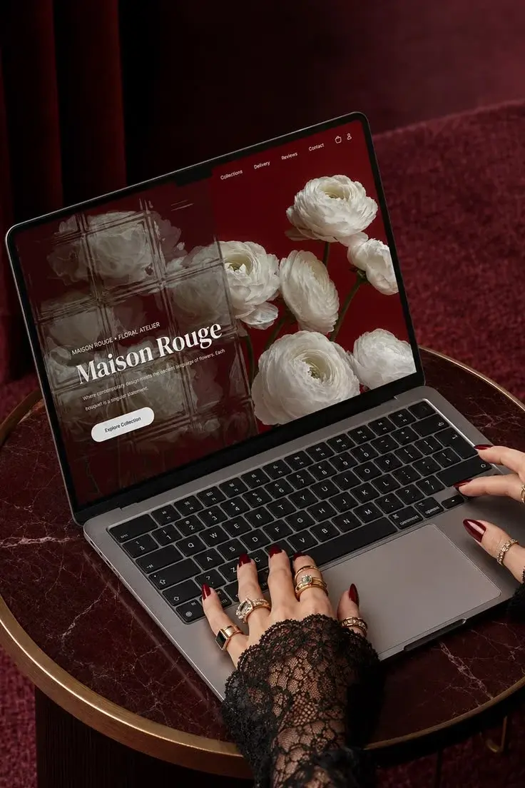 Close-up of hands with lace sleeves typing on a laptop displaying a luxury website titled "Maison Rouge" with white flowers on a red backdrop.