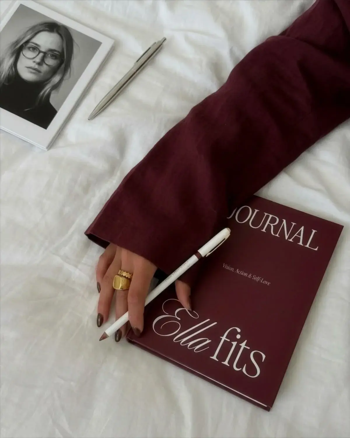 Top-down view of a hand holding a white pen over a burgundy notebook titled "Ella Fits," with a framed portrait nearby.