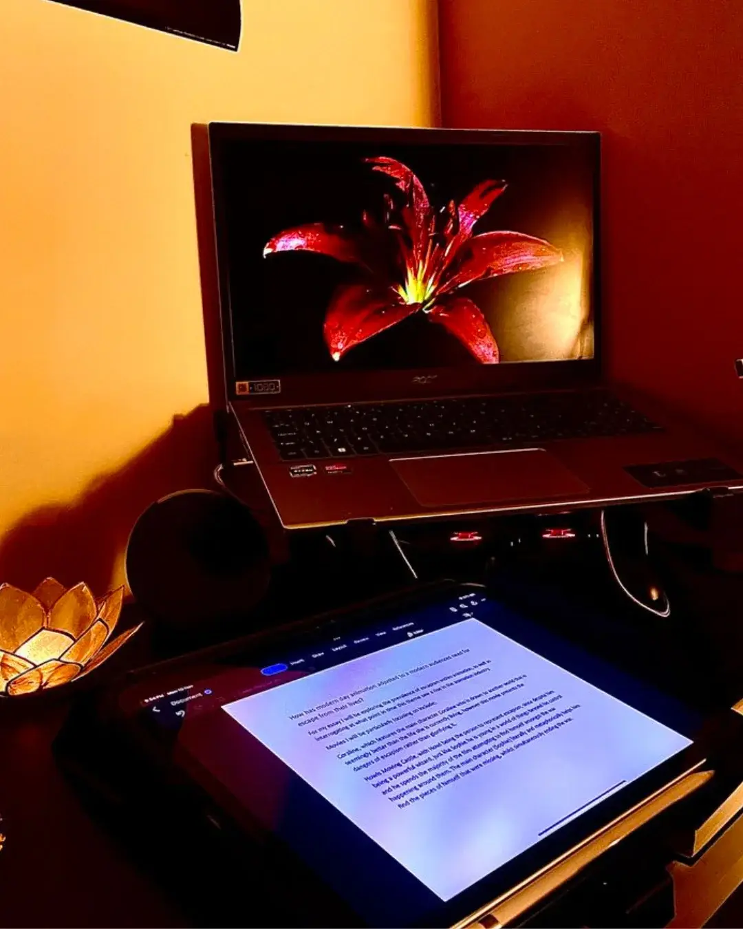 Cozy workspace setup featuring a tablet displaying a document draft alongside a laptop with a red flower wallpaper under warm lighting.